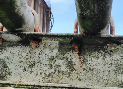 Corroded welded pipe support showing rust and moisture damage under U-bolts